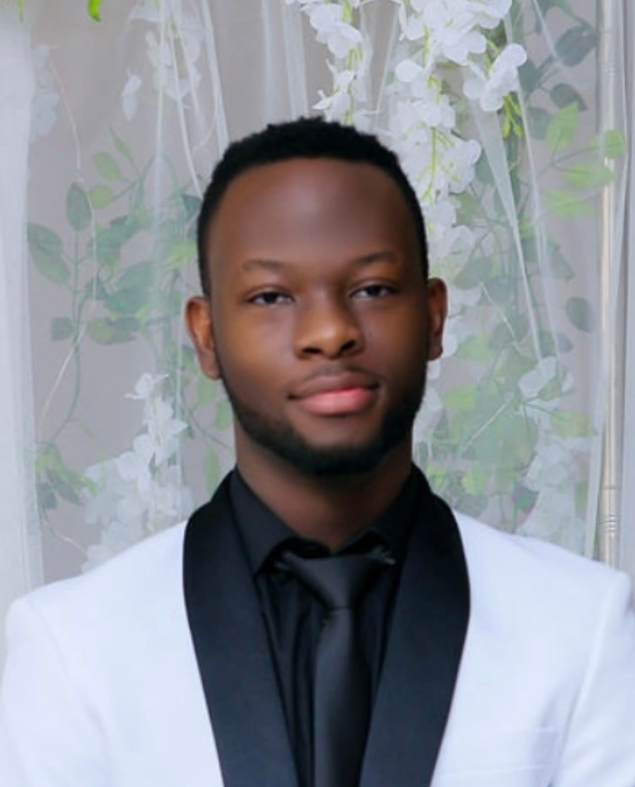 Young Black man in a white tuxedo with black lapels and tie against floral background.