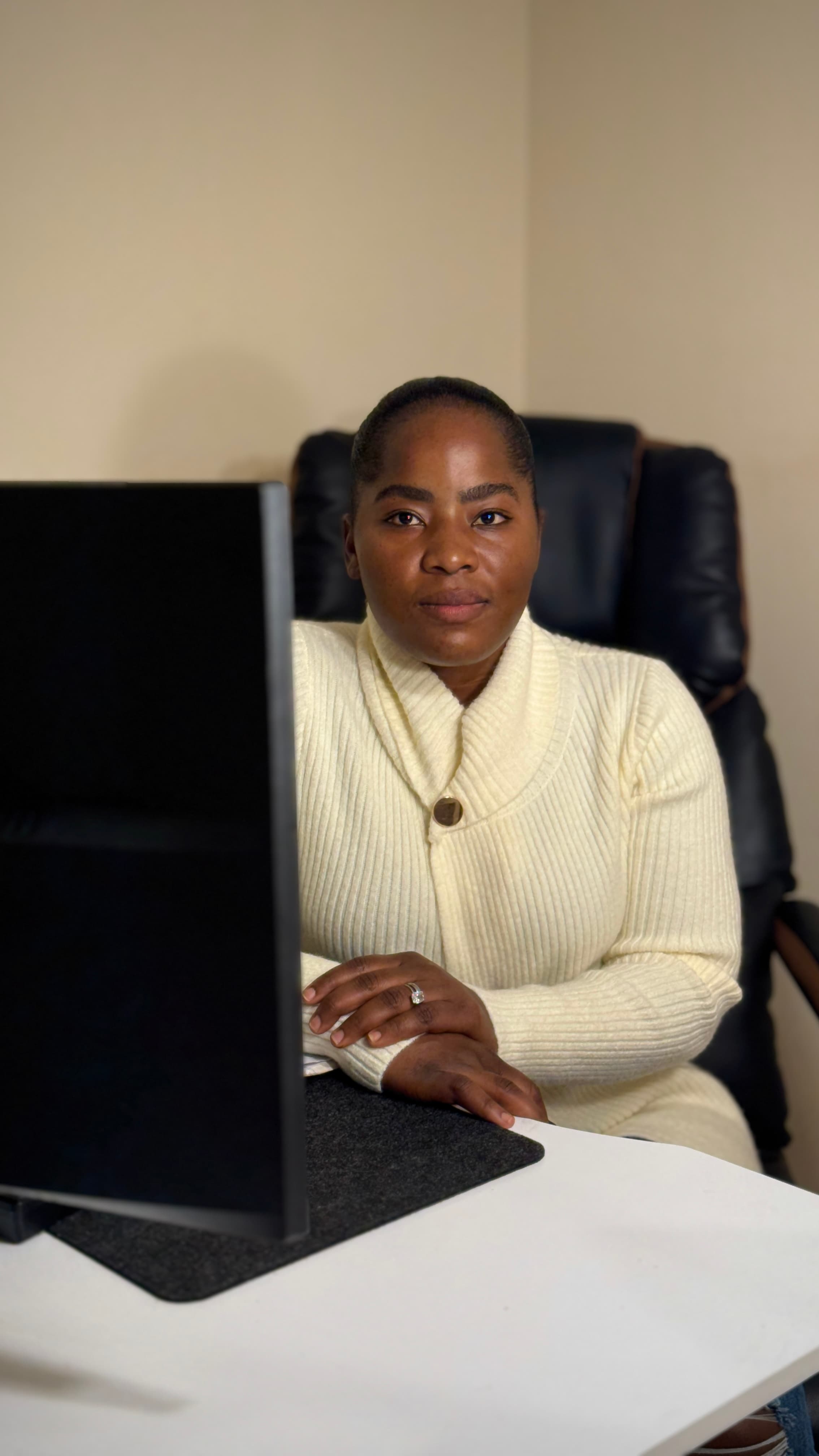 Black woman in a cream sweater sits at a desk behind a monitor, looking forward.